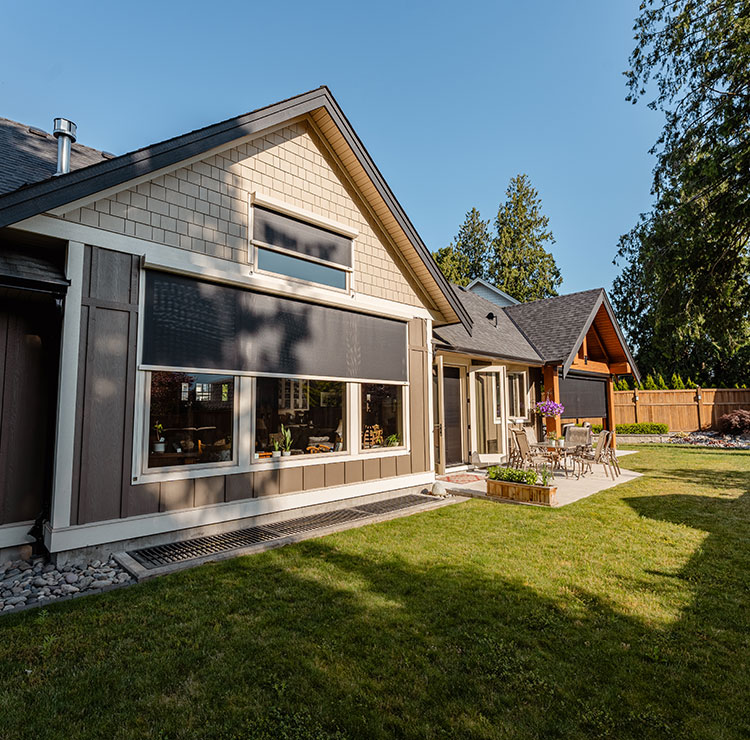 Modern craftsman home exterior with large window featuring exterior solar shade and cable guide system, surrounded by landscaped yard with evergreen trees
