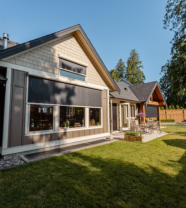 Modern craftsman home exterior with large window featuring exterior solar shade and cable guide system, surrounded by landscaped yard with evergreen trees